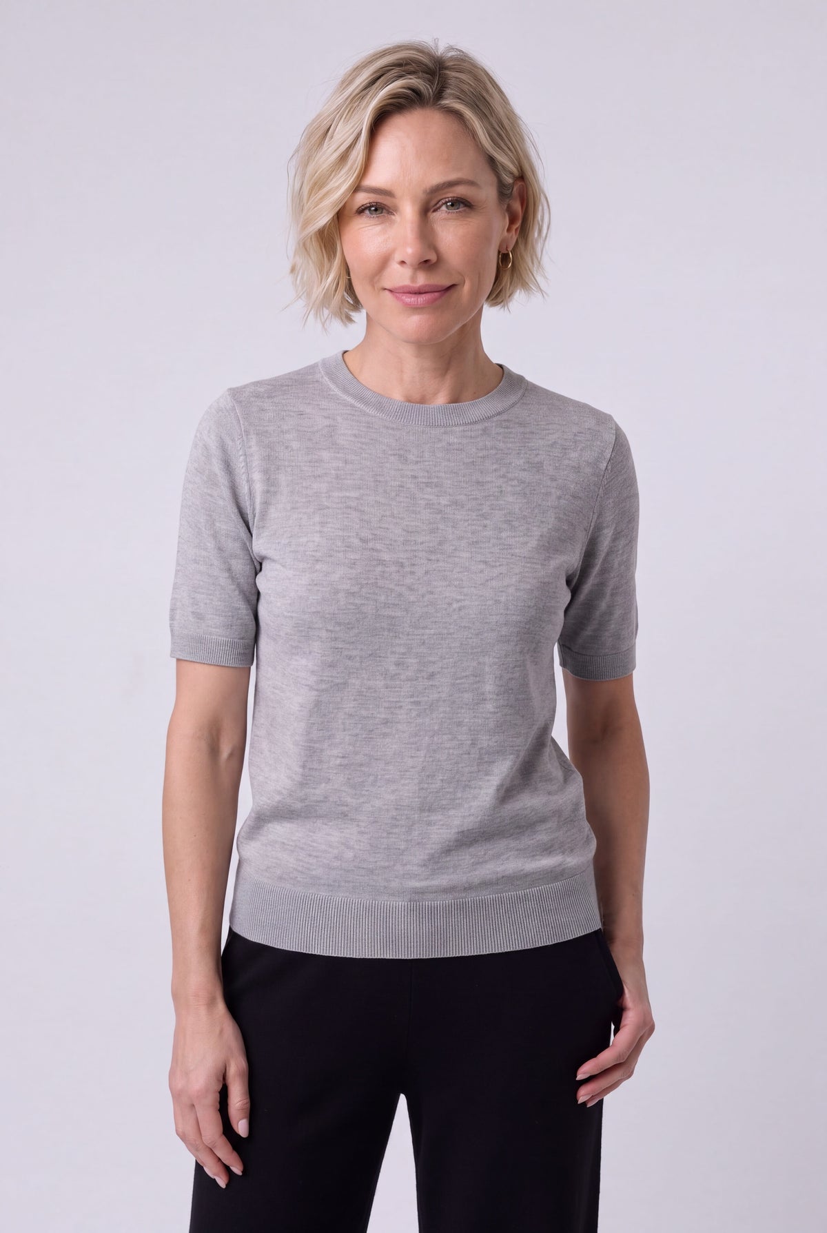 A woman with short blonde hair wearing a light gray short-sleeve crewneck knit sweater and black pants, standing against a light gray studio background.