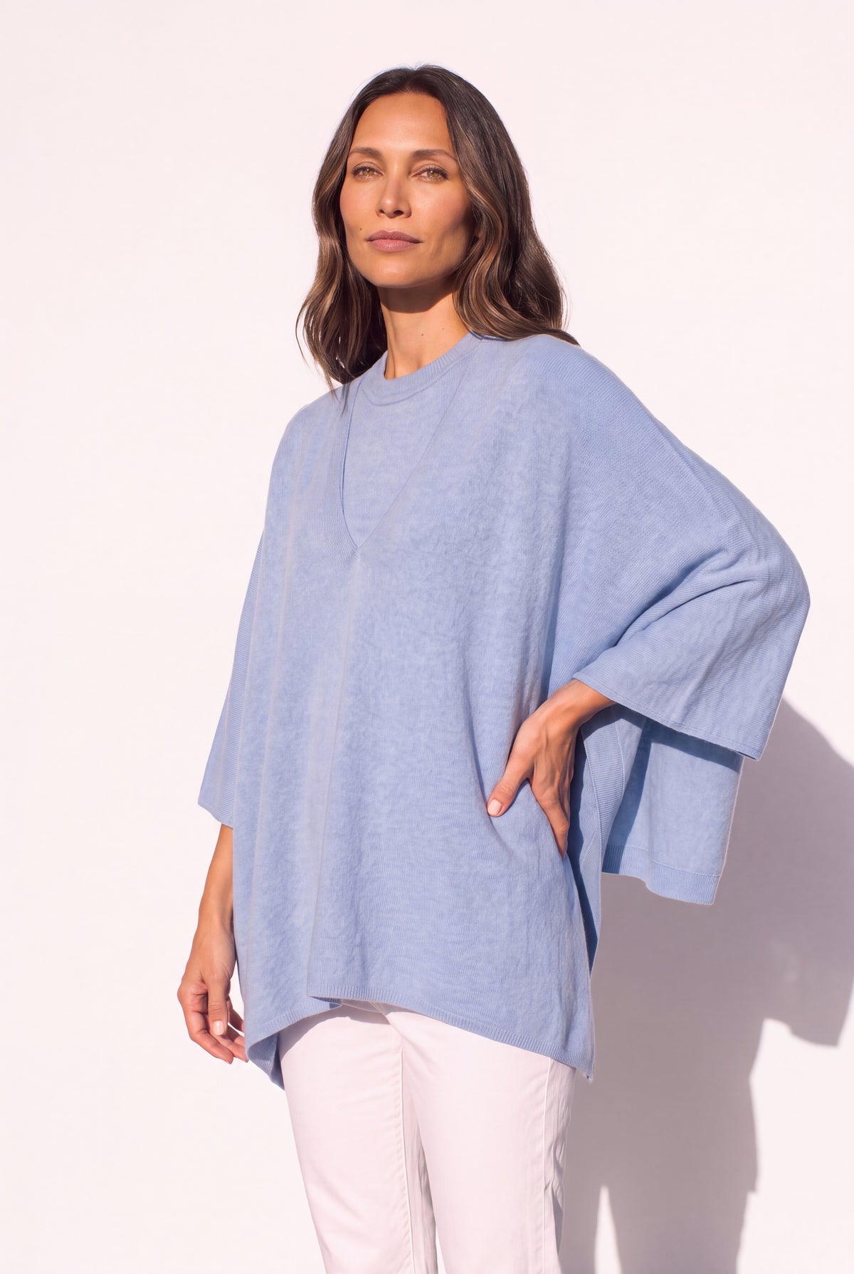 A woman modeling a light blue, oversized poncho-style top with wide three-quarter sleeves and a relaxed silhouette, paired with white pants against a clean white background.