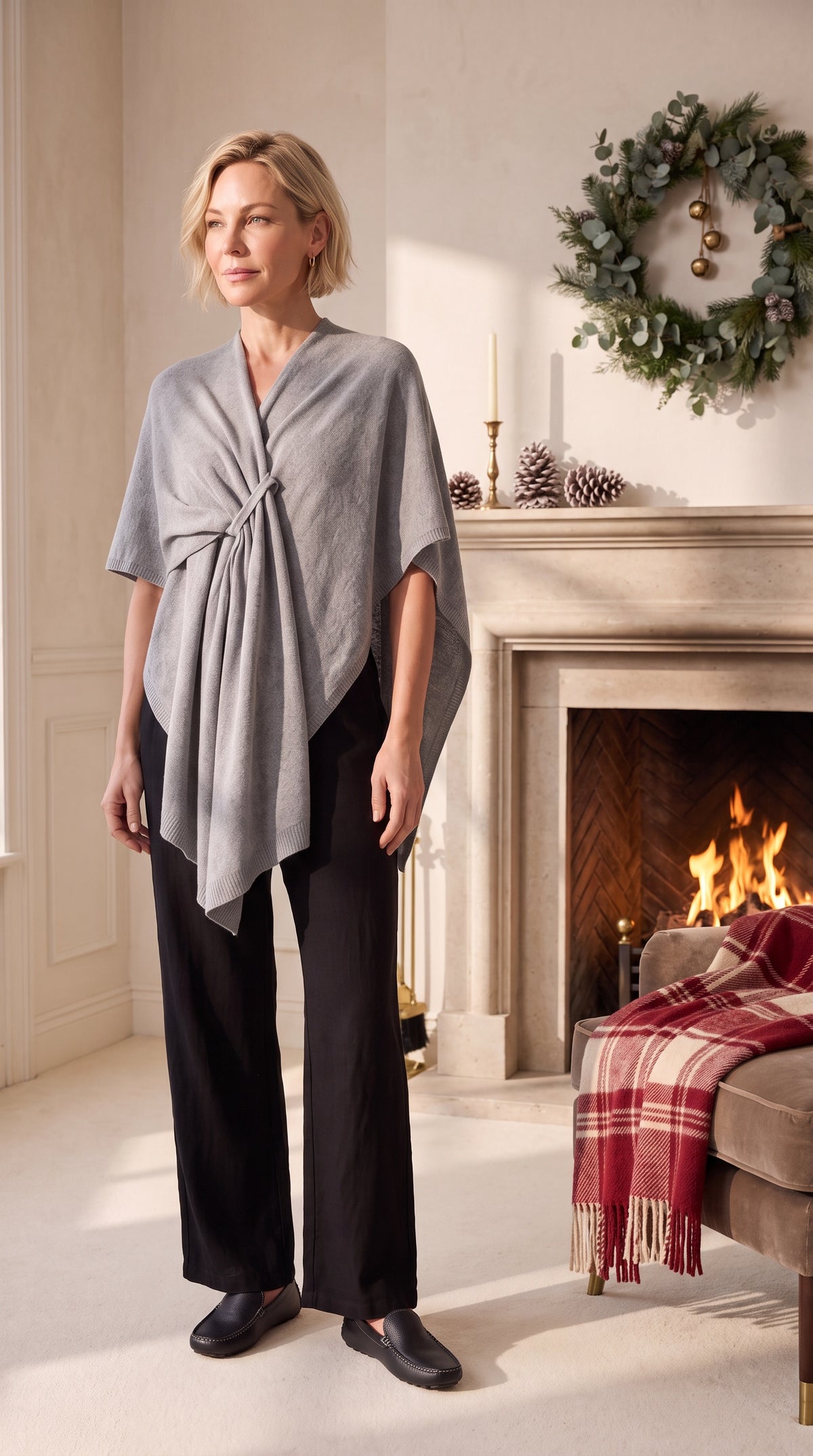 A woman stands in a warmly lit living room wearing a light gray wrap-style top with a twisted front detail and black wide-leg pants. A fireplace with a red plaid throw, a green wreath, and candles are visible in the background.