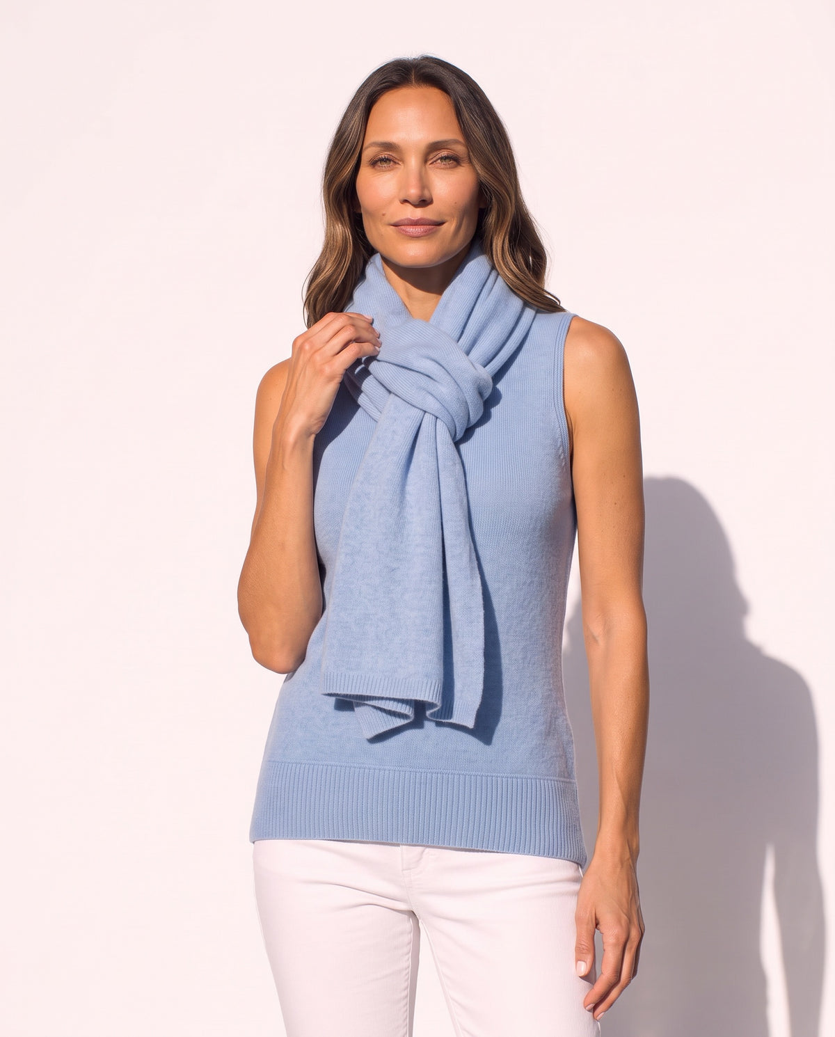 A woman modeling a light blue knit scarf wrapped around her neck, paired with a sleeveless light blue top and white pants, set against a plain light background.