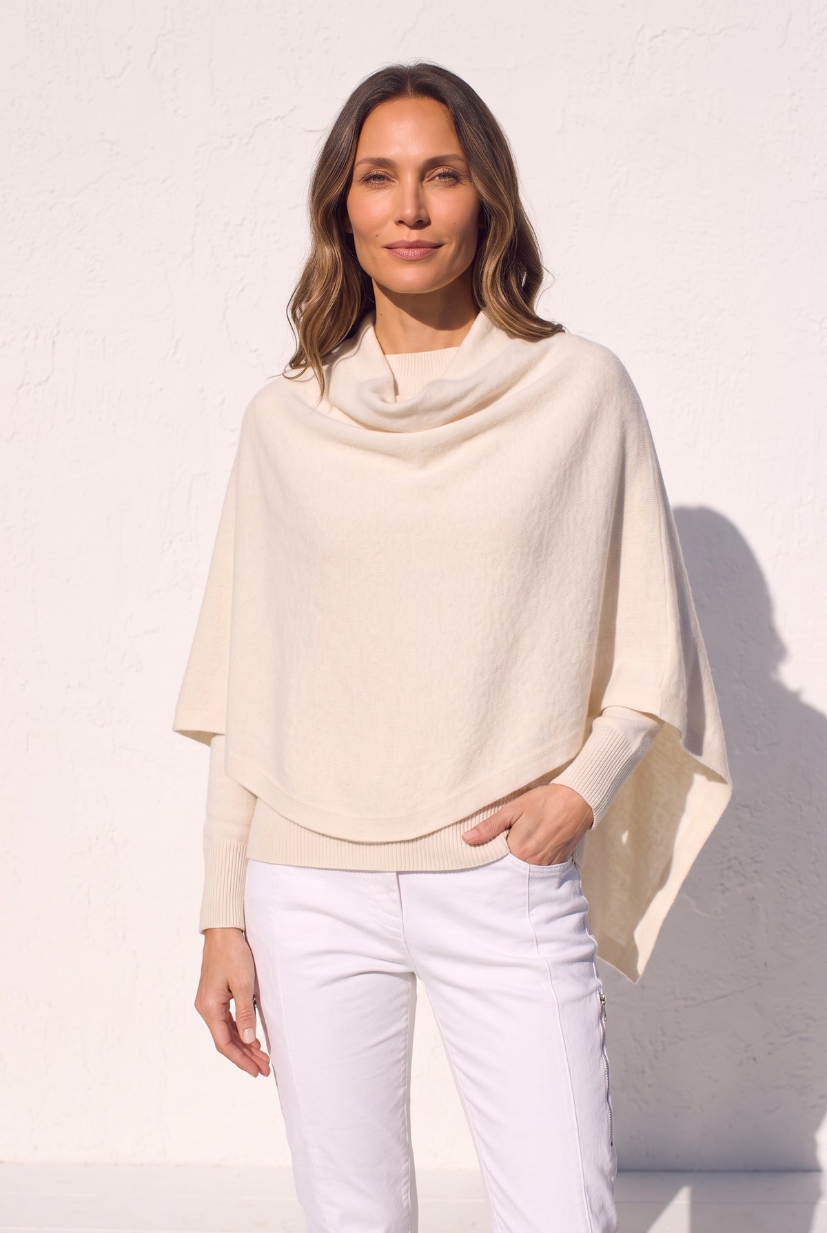 A woman wearing a cream-colored cowl neck poncho sweater and white pants, standing against a white textured wall.