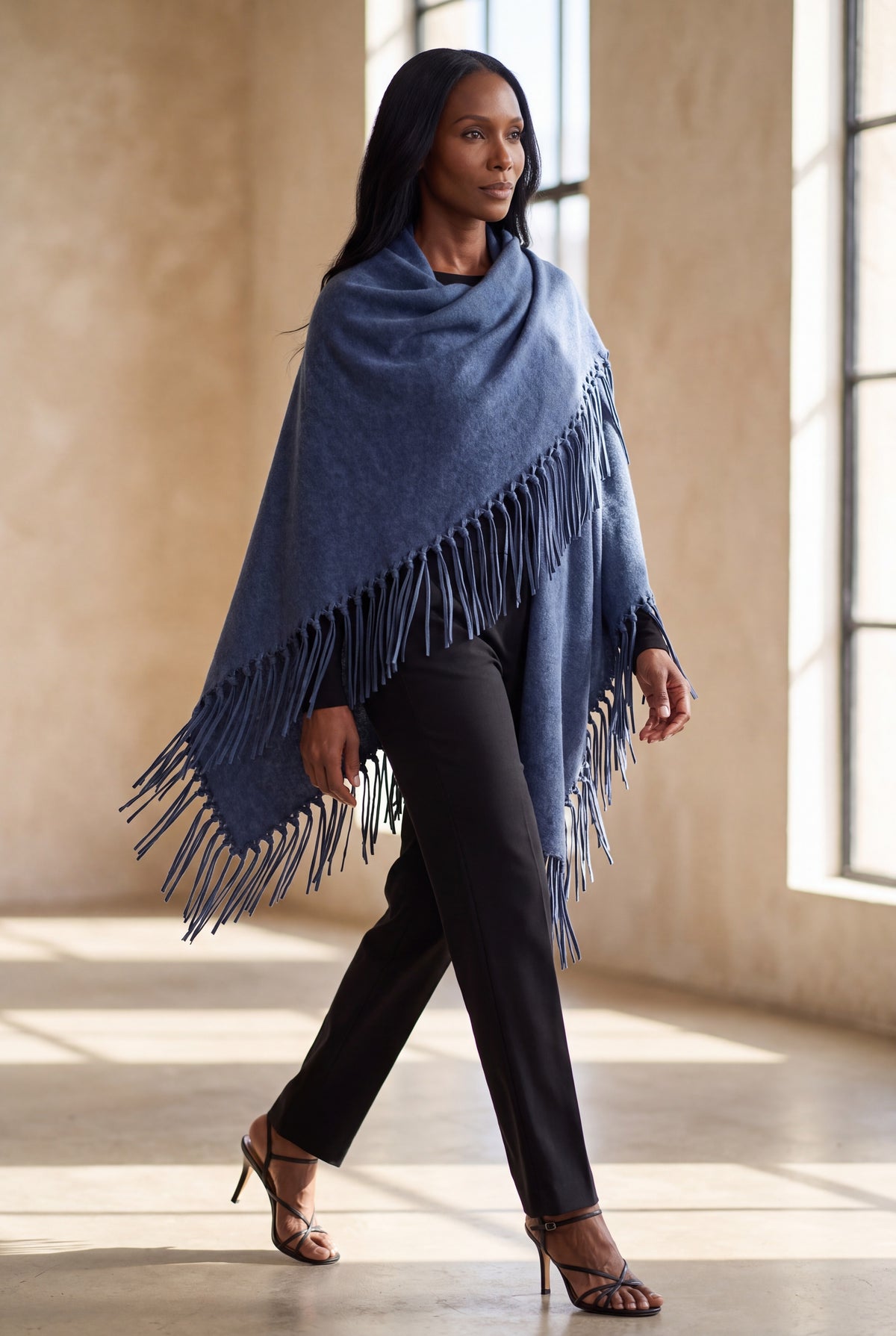 A woman models a blue fringed wrap or poncho draped over her shoulders, paired with black pants and strappy heels in a sunlit indoor space with large windows and textured beige walls.