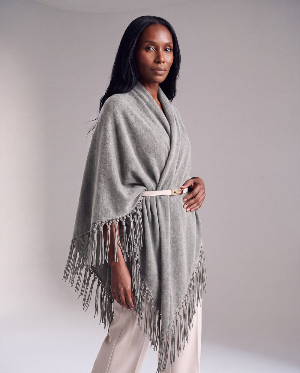 A woman modeling a light gray fringed wrap poncho with a slim white belt, paired with light beige trousers, against a neutral gray studio background.