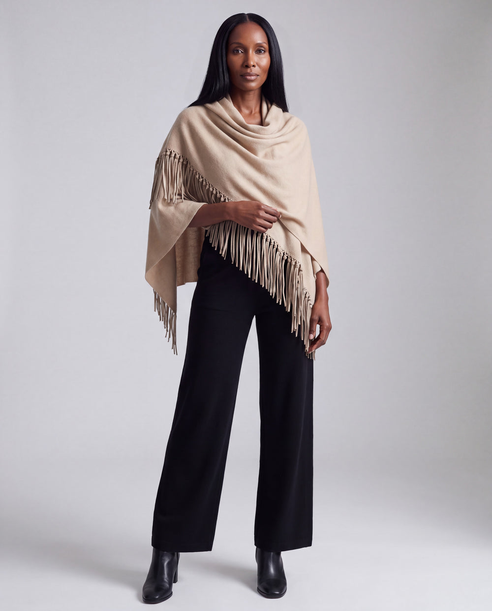 A woman modeling a beige fringed cowl-neck wrap or poncho over black wide-leg pants and black ankle boots, photographed against a light gray studio background.