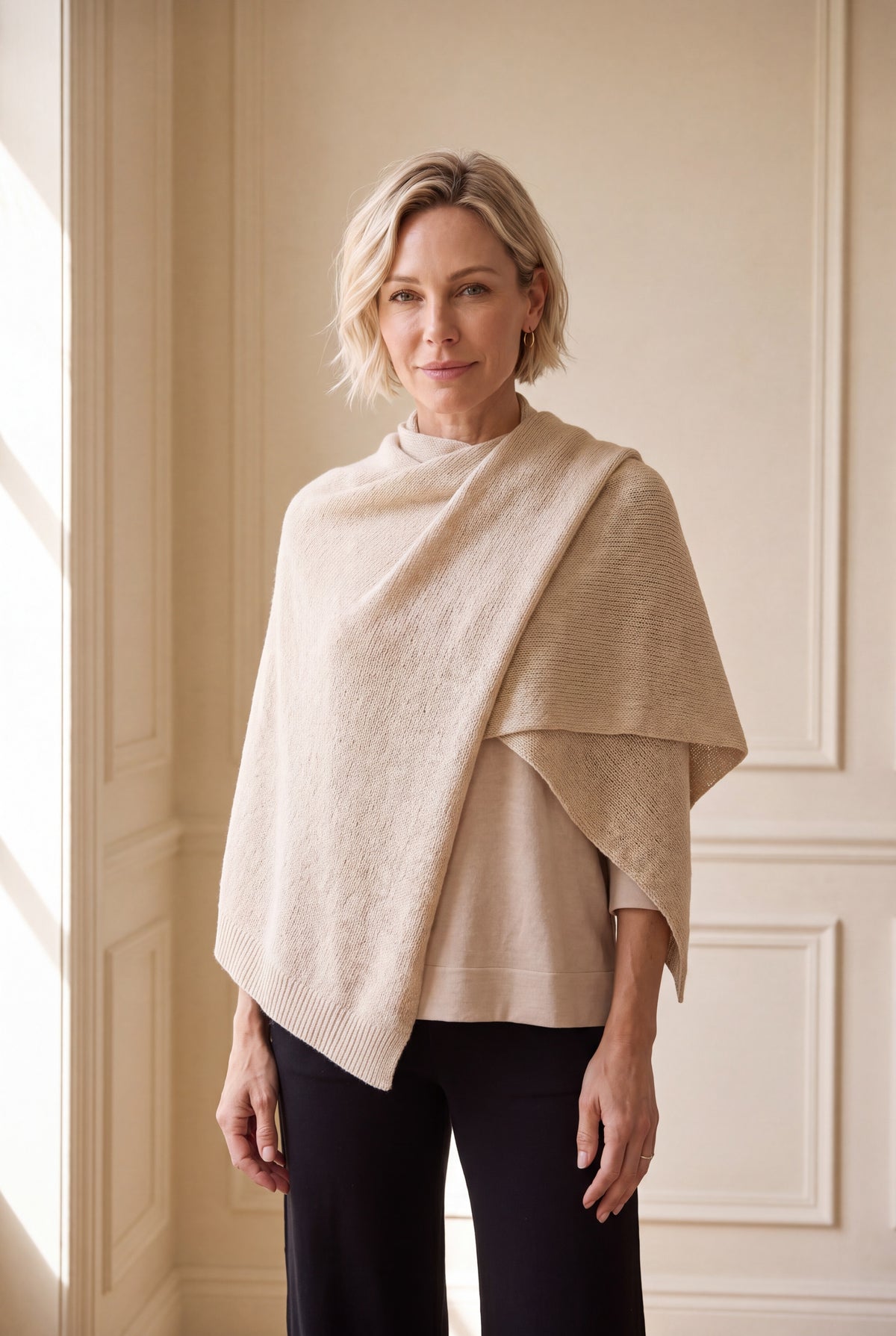 A woman with short blonde hair models a light beige textured knit poncho over a matching beige top and black pants, standing in a brightly lit room with cream-colored paneled walls.