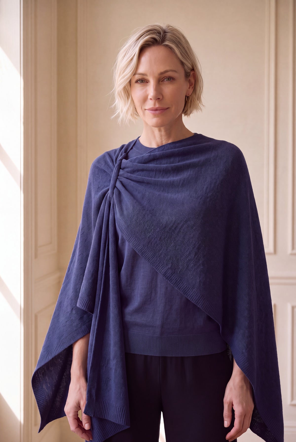 A woman with short blonde hair models a navy blue draped wrap or poncho over a dark top and pants, standing in a brightly lit interior with cream-colored paneled walls.